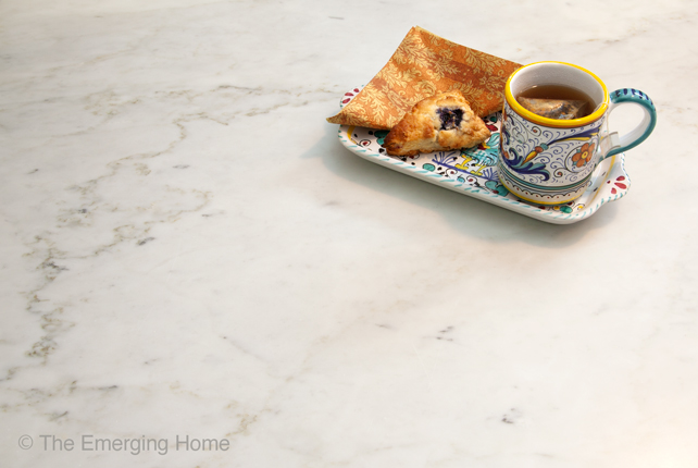 A colorful Italian plate holds a cup of tea and a scone as it sits on a dark veined section of the white marble countertop.