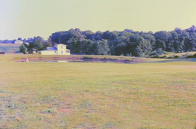 Old white farmhouse in distance with a pond at its side. Large green field in foreground.